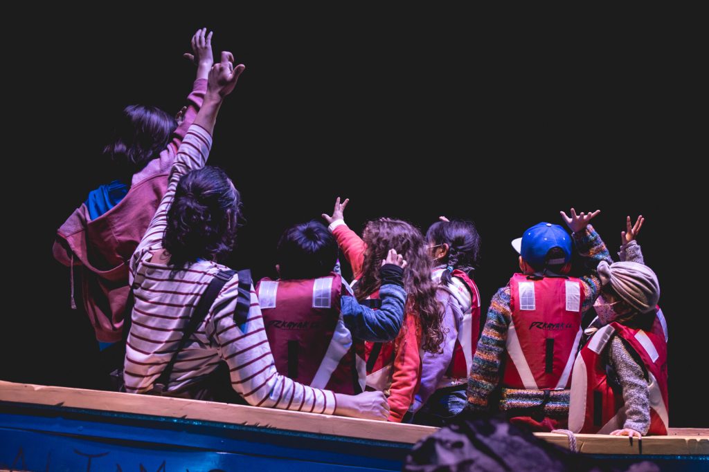Niñas y niños en el público de una obra, levantando las manos
